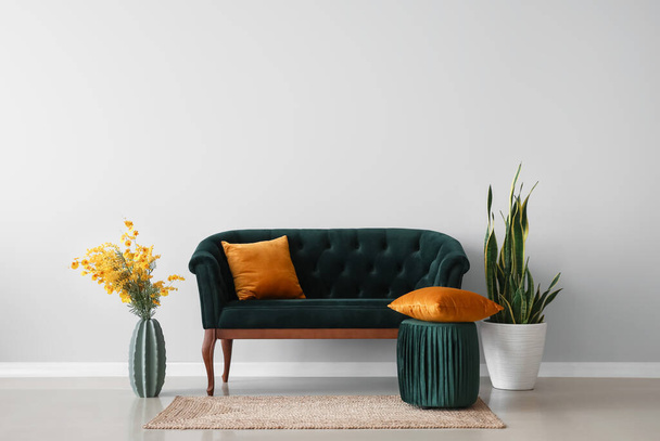Зеленый диван, пуф, ваза с цветами и домашнее растение у светлой стены - Фото, изображение