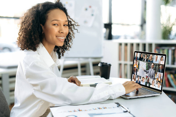 Positive successful young adult African American woman, top manager, head of a company, sitting in front of a laptop in a modern office, conducts online briefing with business partners, taking notes - Foto, imagen