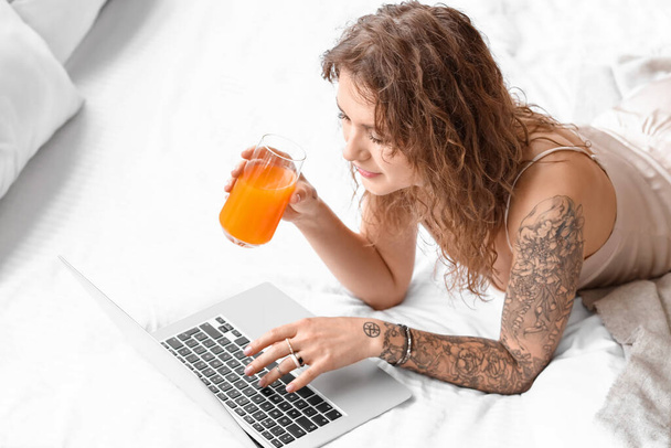 Beautiful tattooed woman with laptop drinking juice in bedroom - 写真・画像