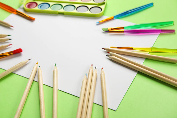 Πολύχρωμα μολύβια, λευκό φύλλο χαρτιού και χρώματα με πινέλα σε πράσινο φόντο - Φωτογραφία, εικόνα