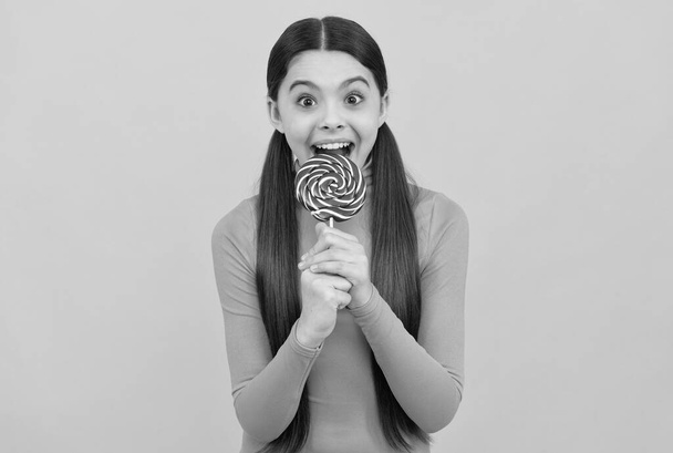 překvapený dospívající dívka kousnutí lízátko cukroví na hůl na žlutém pozadí, zubní péče - Fotografie, Obrázek
