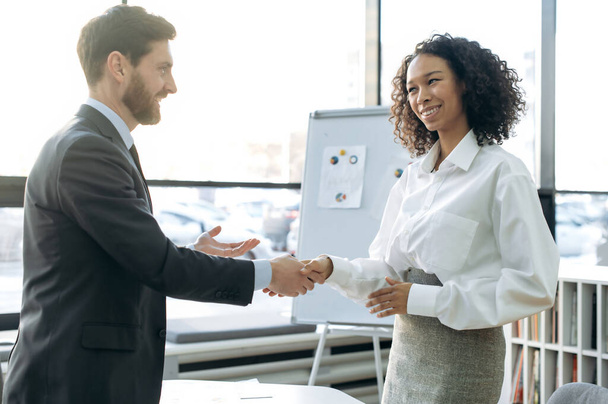 Business partners, man and woman of different nationalities, stand in modern office, hold business negotiations, made a good deal, agreed on cooperation, greet each other shaking hands, smiling - Фото, изображение
