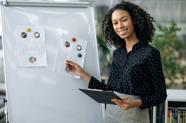 Purposeful successful African-American young woman, in formal clothes, financial manager, economist, trader, stands in an office near a white board, presents her report of project, smiling friendly - Foto, afbeelding