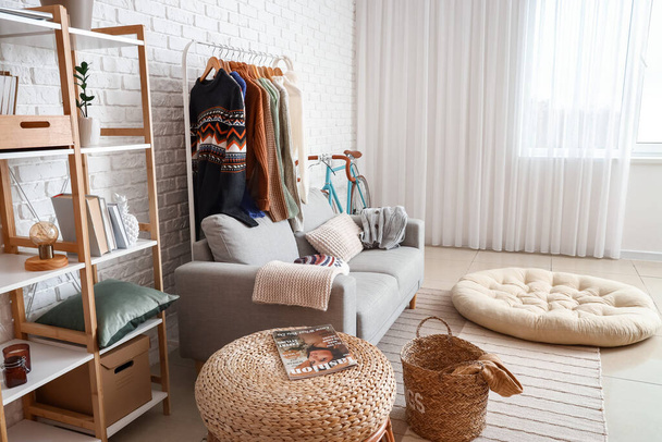 Sofa, shelf unit and rack with sweaters near white brick wall in living room - 写真・画像