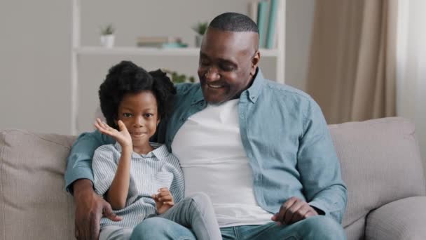 Afričanky americká rodina sedí na pohovce dítě dívka mává ahoj pozdrav dospělý otec objímání dcera mluví na kameru s úsměvem ukazující zdravé bílé zuby po zubní procedury video chat webcam pohled - Záběry, video