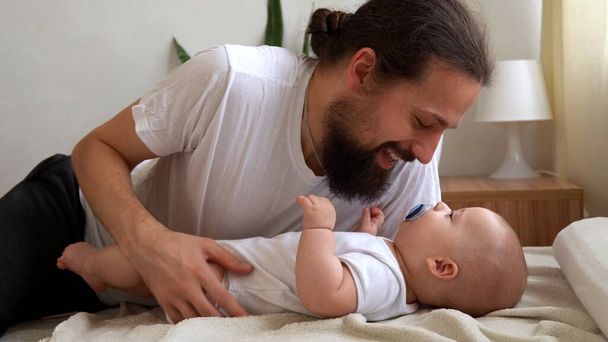 Authentic Young Bearded Man Holding Newborn Baby. Dad And Child Son On Bed. Close-up Portrait of Smiling Family With Infant On Hands. Happy Marriage Couple On Background. Childhood, Parenthood Concept - Fotografie, Obrázek
