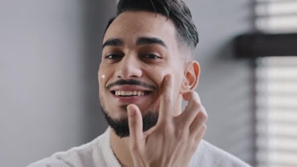 Close-up male happy face with perfect skin mirror view hispanic arab man bearded handsome healthy guy smiling smearing cheeks with moisturizing cream natural anti wrinkle gel after shave beauty ritual - Footage, Video