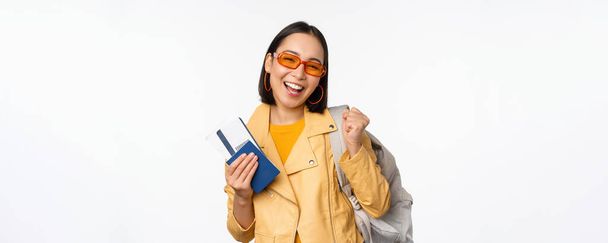 Happy asian girl going on vacation, holding passport and flight tickets, backpack on shoulder. Young woman tourist travelling abroad, standing over white background - Fotó, kép