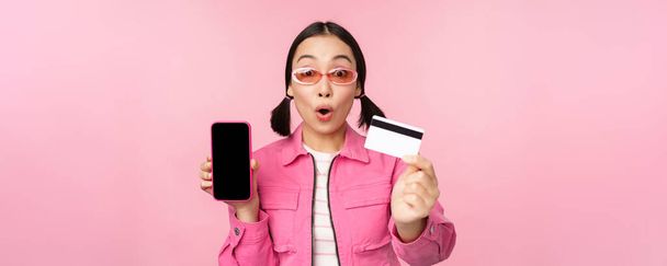 Азиатская девушка показывает экран мобильного телефона и кредитной карты, реагирует удивленно на камеру, задыхаясь впечатлен, стоя на розовом фоне, концепция покупок в Интернете - Фото, изображение