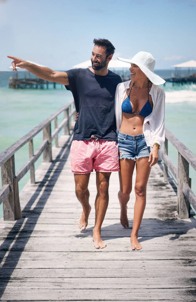 Gehen wir mal rüber. Ganzkörperaufnahme eines glücklichen jungen Paares, das Arm in Arm während eines gemeinsamen Urlaubs eine Promenade hinunterläuft. - Foto, Bild