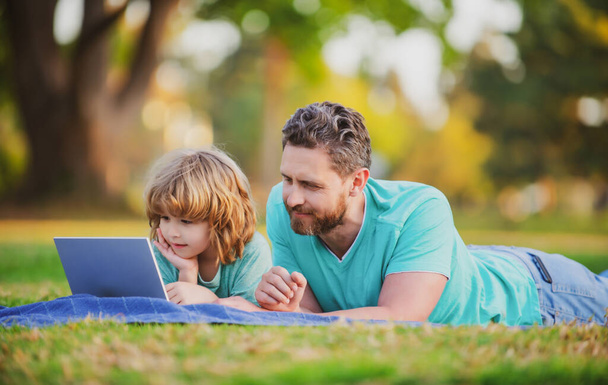 Szczęśliwy ojciec za pomocą laptopa zrelaksować się z uczniem syn gospodarstwa laptopa bawić się razem, uśmiechnięty tata i mały chłopiec dziecko cieszyć weekend z gadżetów na zewnątrz na łonie natury. - Zdjęcie, obraz