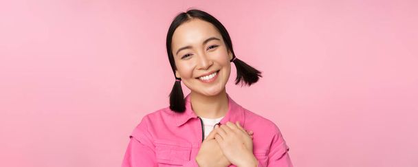 Schattig koreaans meisje met domme haarballen, op zoek gelukkig en dankbaar, dank u pose, hand in hand op hart gevleid, poseren tegen roze achtergrond - Foto, afbeelding