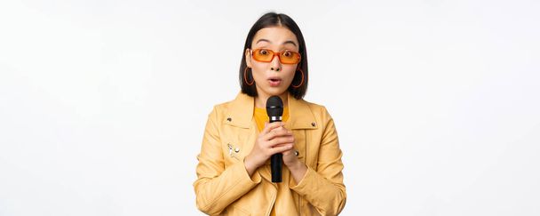 Portrait of beautiful asian woman in sunglasses, stylish girl singing, giving speech with microphone, holding mic and smiling, standing over white background - Photo, image