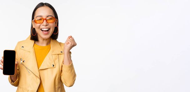 Щаслива азіатська дівчина в сонцезахисних окулярах, показує екран мобільного телефону, інтерфейс смартфона, сміється і посміхається, святкує, стоїть на білому тлі
 - Фото, зображення