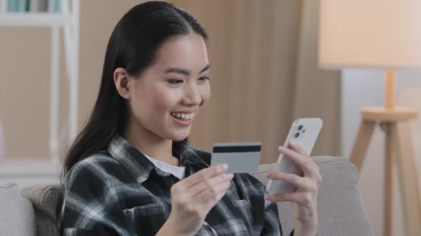 Χαμογελώντας ασιατική εθνοτική γυναίκα πελάτης χρήστη κρατώντας τραπεζική πιστωτική κάρτα με εικονικά χρήματα πληκτρολογώντας smartphone στο σπίτι. Ευτυχής πολυεθνική θηλυκό αγοραστής χρησιμοποιώντας εύκολη εφαρμογή κινητών πληρωμών κάνοντας online αγορά - Πλάνα, βίντεο