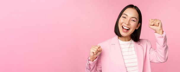 Enthousiaste aziatische corporate vrouw, zakenvrouw heffen hand omhoog en juichen, triomferen, winnen en vieren, staan over roze achtergrond - Foto, afbeelding