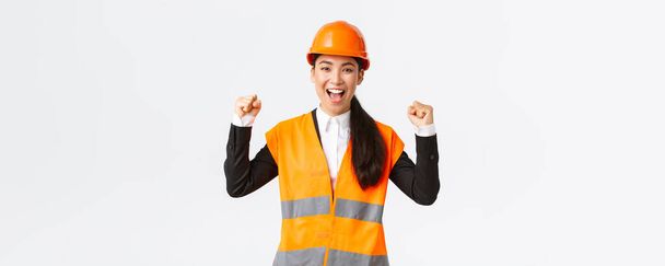Successful winning female asian engineer fist pump and shout yes delighted, wear safety helmet and jacket, triumphing over victory, finish building in time, standing satisfied white background - Photo, Image