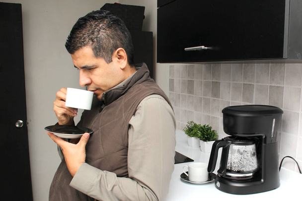 Dark-haired Latino adult man prepares a cup of coffee in a coffee maker to smell and taste at breakfast in the kitchen is thoughtful, worried, thoughtful, depressed - Photo, Image