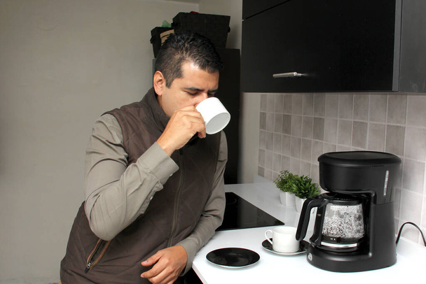 Koyu renk saçlı, yetişkin bir adam kahve makinesinde kahvesini koklamak ve tatmak için hazırlar. Mutfakta bu düşünceli, endişeli, düşünceli ve depresiftir. - Fotoğraf, Görsel