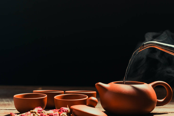 Ρίχνουμε ζεστό νερό σε μια πήλινη κανάτα τσαγιού για αρωματικό κόκκινο τριαντάφυλλο τσάι. Τοποθετείται σε παλιό ξύλο με μαύρο φόντο - Φωτογραφία, εικόνα