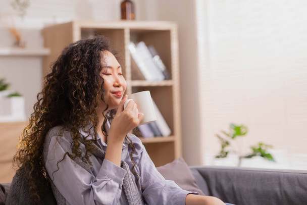 Asian Woman pondering at home in her leisure time while reclining on the sofa and drinking hot tea. On a lovely winter morning, a joyful girl relaxes at home. - Photo, image