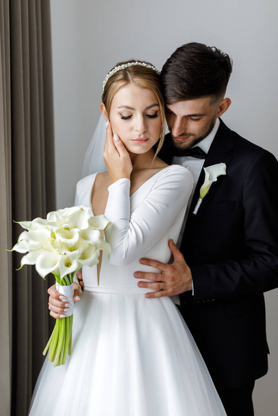 A stylish bridegroom cute embraces a bride in a bridal dress. The newlyweds embrace in the room and spend time together - Foto, afbeelding