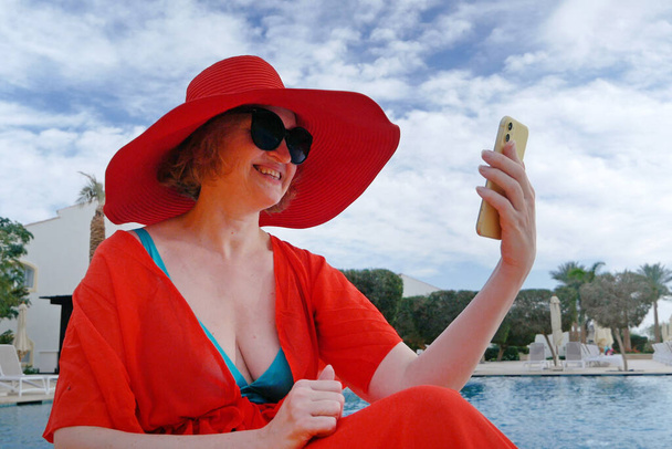 Letnie wakacje kobieta w czerwonym kapeluszu i peleryna na plaży z telefonem komórkowym w pobliżu basenu pływać amd palmy, kobieta blogger, na zewnątrz - Zdjęcie, obraz