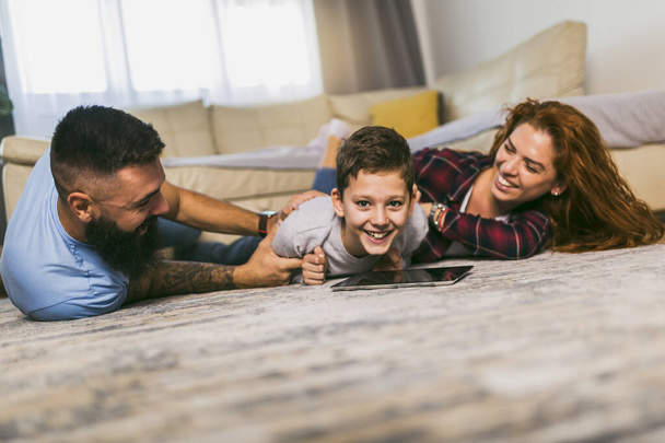 Счастливая семья с одним ребенком, лежащим на ковре и пользующимся цифровым планшетом
 - Фото, изображение