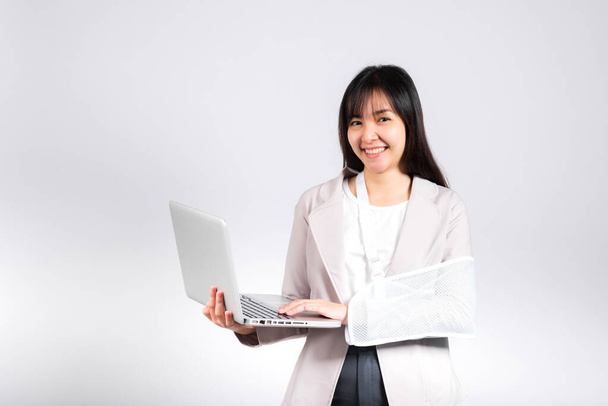 Asian business woman confident smiling broken arm after accident and wear arm splint for treatment but still have to work she hold laptop computer studio isolated on white background,copy space - Фото, зображення