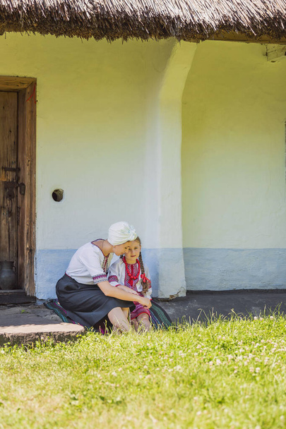 mother and daughter in Ukrainian national costumes are sitting near the house - 写真・画像