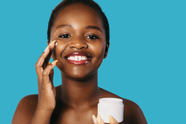 アフリカ系アメリカ人女性の笑顔は、青い背景に隔離されたポーズで、彼女の顔に美容製品を適用します。孤立した背景。美しい黒人女性だ。スキンケア. - 写真・画像