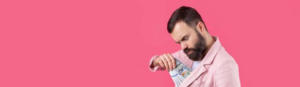 Ritratto di un giovane uomo d'affari soddisfatto con la barba vestita con una giacca rosa che ci mostra banconote in dollari su uno sfondo rosso da studio. Gusto, odore di soldi. - Foto, immagini