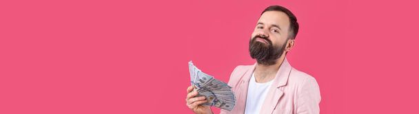 Portrait of a contented young businessman with a beard dressed in a pink jacket showing us dollar banknotes against a red studio background. Taste, smell of money. - Foto, imagen