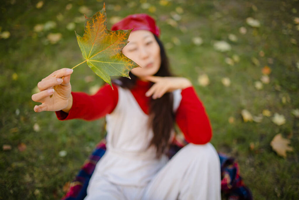 Syksyn mieliala. Kaunis nuori aasialainen nainen nahkahatussa makaa syksyllä ruohoa ja keltaisia lehtiä. Kausiluonteinen syksyn muoti. - Valokuva, kuva
