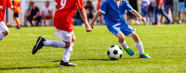 Boys play soccer. Teenagers playing soccer in sports field. Children kicking soccer game outdoors. Young male footballers running in red and blue sports uniforms - 写真・画像