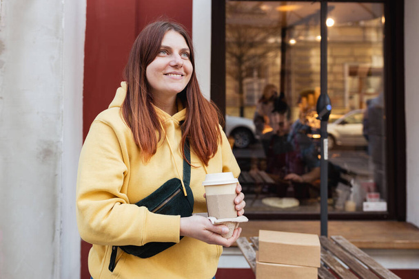 Young stylish hipster redhead woman walking on street, wearing trendy outfit and drinking hot latte. Urban lifestyle banner. Traveler. Take away delivery concept. Eco recycle environment friendly. - Fotó, kép