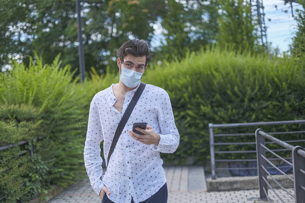 όμορφο Ιταλό μελαχρινό αγόρι με μάσκα για τον ιό covid στο τηλέφωνο. Υψηλής ποιότητας φωτογραφία - Φωτογραφία, εικόνα