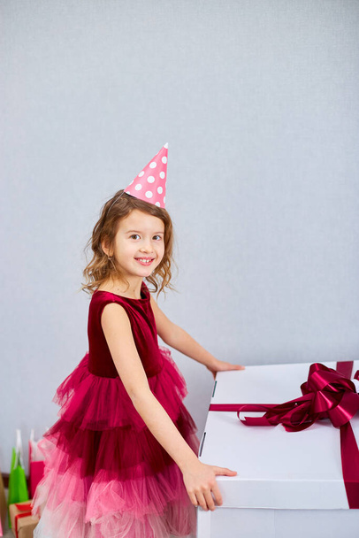 Radosna dziewczynka w różowej sukience i kapeluszu otworzyć duży prezent pudełko z balonami w domu urodzinowe serpentyny, Wszystkiego najlepszego. Świętowanie - Zdjęcie, obraz