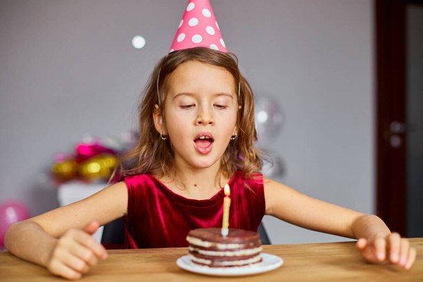 Mała dziewczynka ma na sobie urodzinowy kapelusz patrząc na tort urodzinowy i dmuchanie świec na przyjęcie koncepcyjne uroczystości. - Zdjęcie, obraz