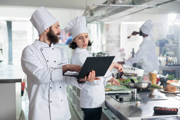 Head chef with modern laptop showing gourmet dish recipe to sous chef required for gastronomic event held at restaurant. - Фото, изображение