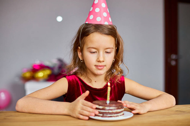 Mała dziewczynka nosi kapelusz urodzinowy myśli życzenie, patrząc na tort urodzinowy, z świecącymi świecami dla świętowania koncepcji party. - Zdjęcie, obraz