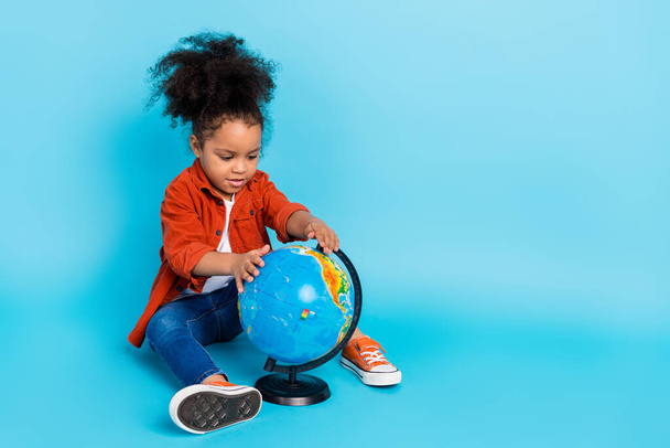 Full length photo of funky curly little girl sit with globe wear shirt jeans shoes isolated on blue background - Foto, Bild