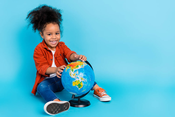 Full body photo of impressed wavy small girl sit with globe wear shirt jeans sneakers isolated on blue background - Foto, Bild