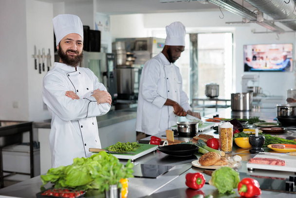Usmívající se sebevědomý šéfkuchař stojící v restauraci profesionální kuchyně při pohledu na kameru po přípravě receptu. - Fotografie, Obrázek