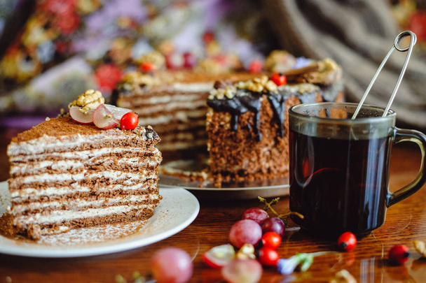 a piece of cake Medovika on a white plate, decorated with nuts, grapes. Honey cake with creamy cream on a wooden table, a mug of tea. - 写真・画像