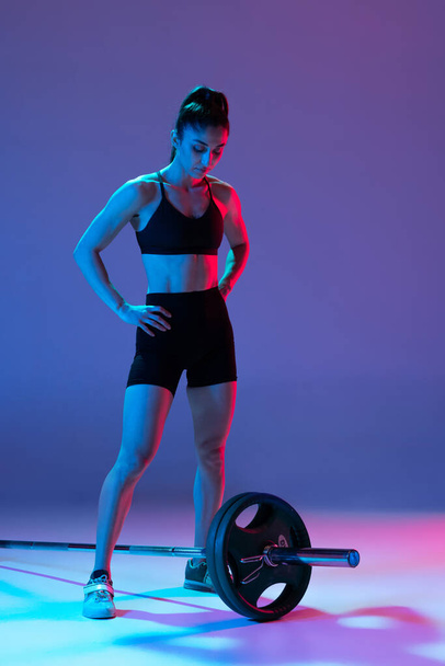 Studio shot of female athlete, sportive woman posing with barbell isolated on purple background in neon light. Sport, beauty, strength concept - Φωτογραφία, εικόνα
