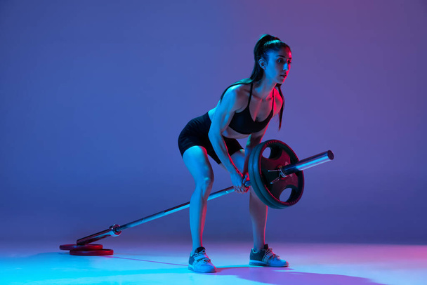 Studio shot of female athlete, sportive woman posing with barbell isolated on purple background in neon light. Sport, beauty, strength concept - Foto, Imagem