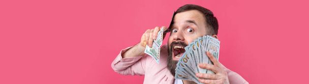 Portrait d'un jeune homme d'affaires heureux avec une barbe vêtue d'une veste rose nous montrant des billets en dollars sur fond de studio rouge. Goût, odeur d'argent. - Photo, image