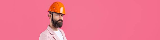 Portret młodego inżyniera budowlanego nosi pomarańczowy twardy kapelusz, w różowej kurtce stojącej na czerwonym tle pracowni. - Zdjęcie, obraz