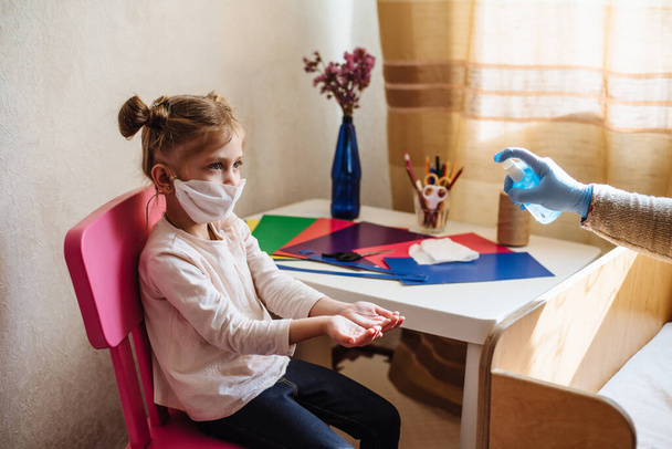 Mom sprays a disinfectant antibacterial spray on the girl's hands. Preventive measures against Covid-19 infection. Sick child in protective mask on isolation at home. - Фото, изображение